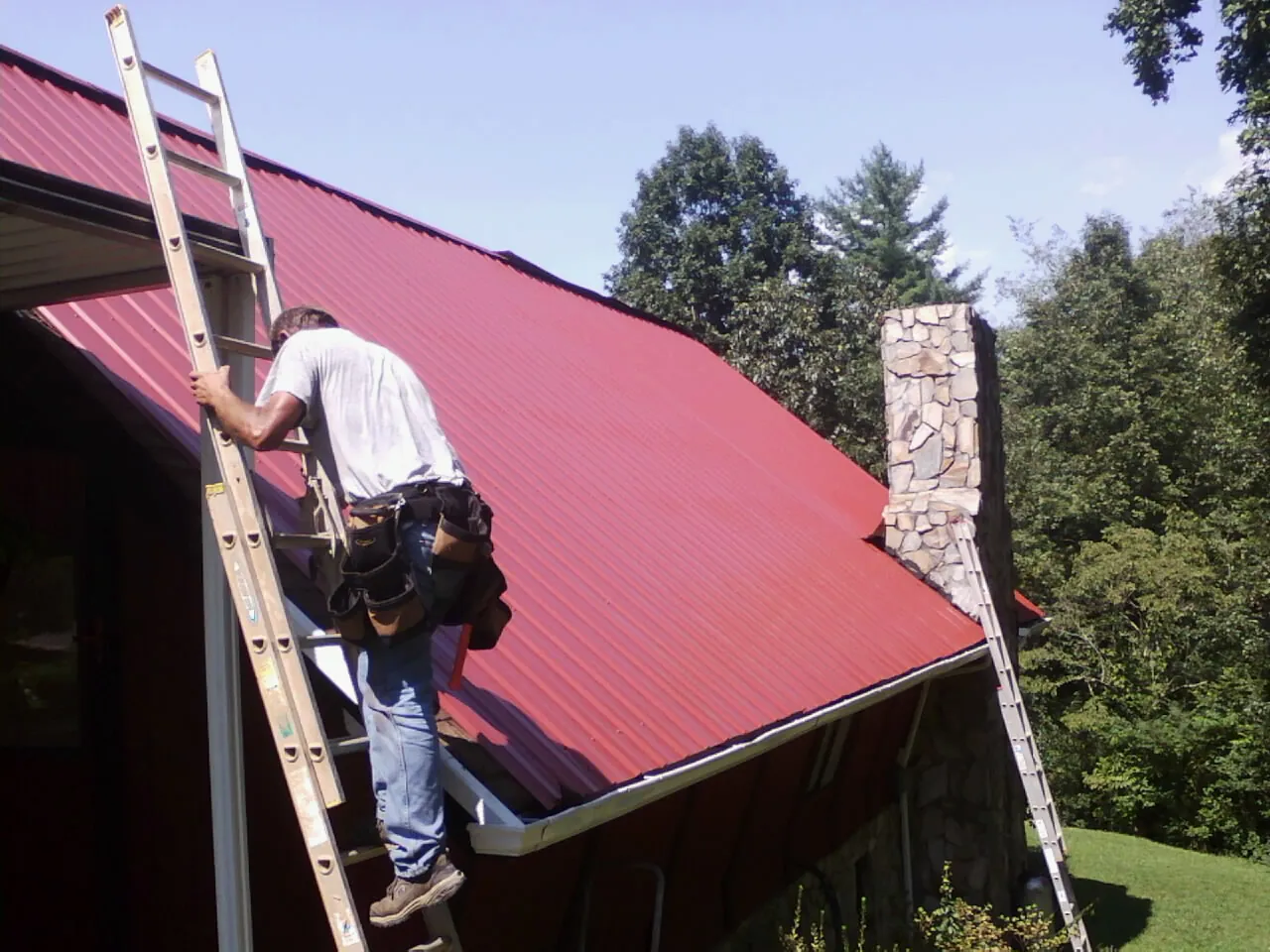 Roofer on a ladder beside a red metal roof and stone chimney — Soffit & Fascia Repair by Roofing Brothers in Georgetown